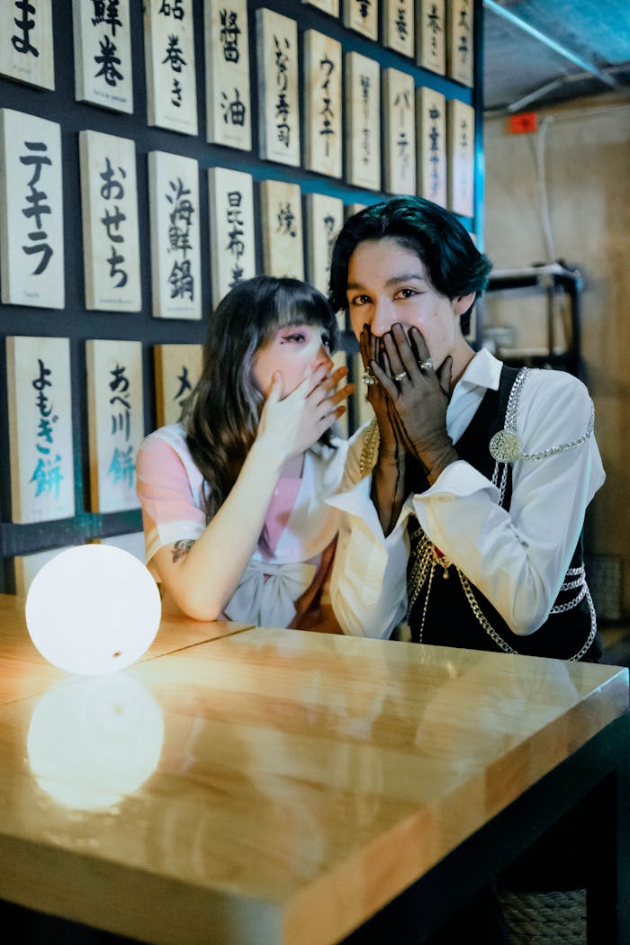 Two friends whisper at a table in a Japanese-themed café with ambient lighting.