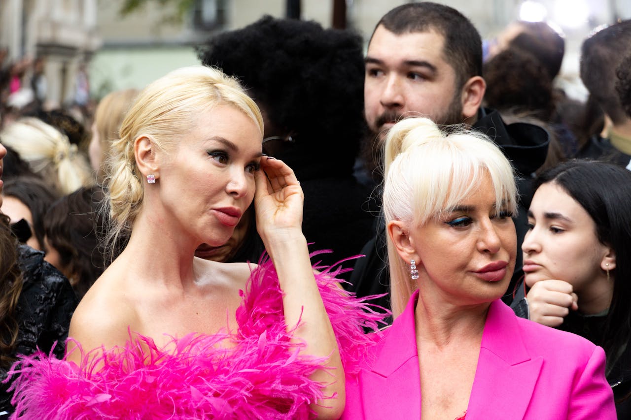 Stylish women in glamorous pink outfits amidst a lively crowd, showcasing fashion and celebration.