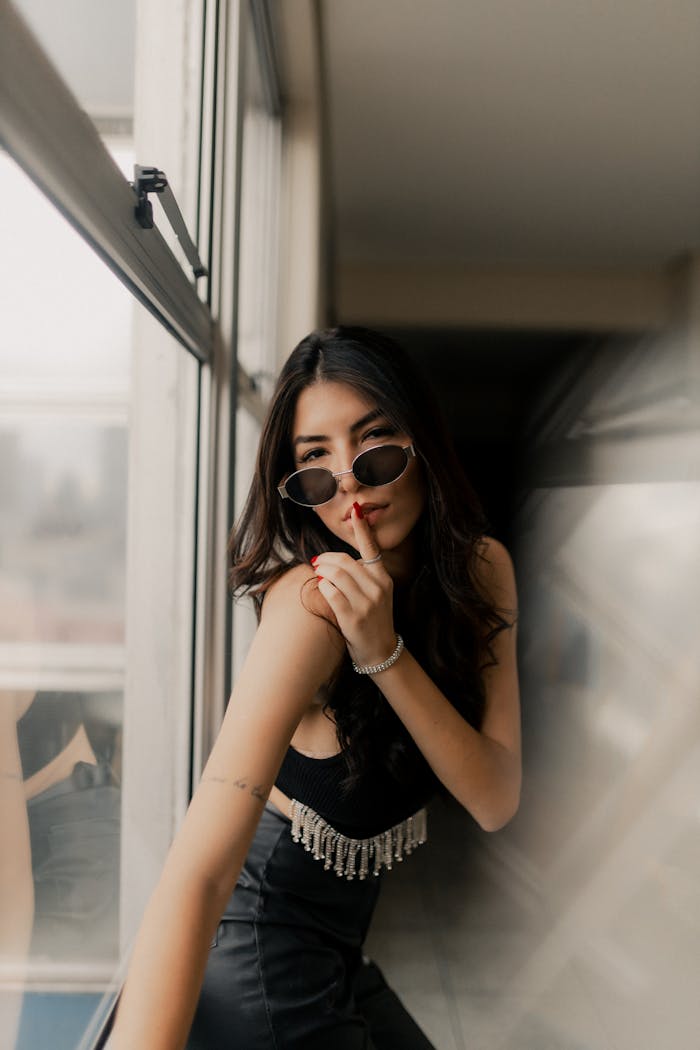 Stylish woman in sunglasses poses by a window indoors, exuding elegance and mystery.