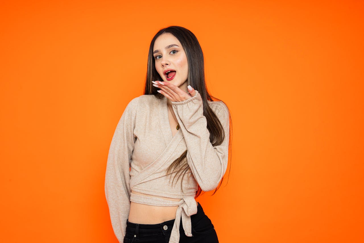 Stylish young woman in trendy outfit posing with playful expression on bright orange background.