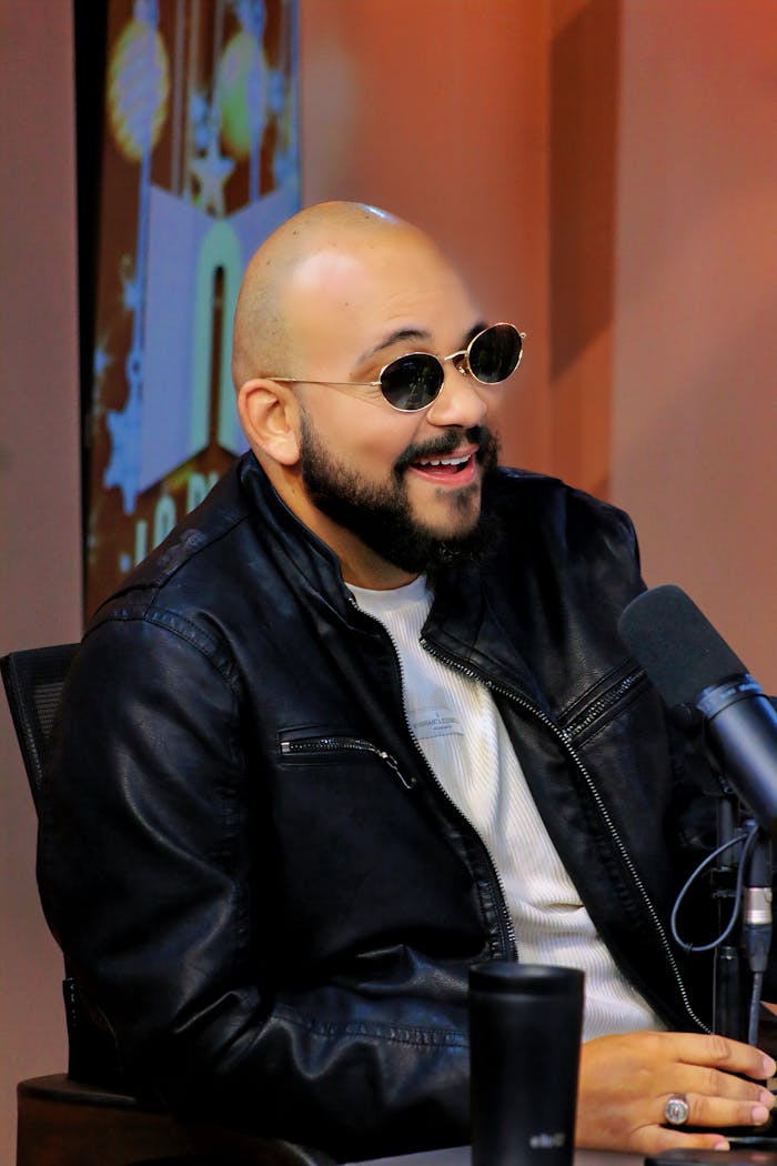 Bald man with sunglasses in a leather jacket speaking into a microphone at an event.
